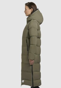 Lang olivengrøn pufferjakke med høj krave, sidelommer med lynlås og hætte. Har et quiltet design med en glat tekstur.