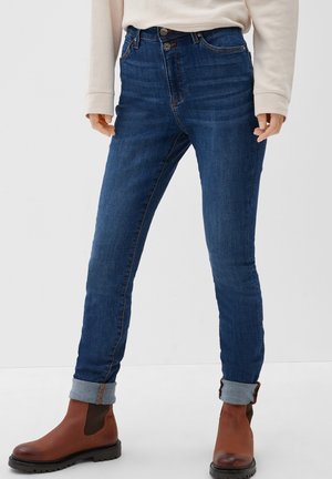 Person wearing dark blue rolled-up skinny jeans, beige long-sleeve top, and brown leather ankle boots standing on a white floor.