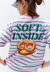 Gestreepte t-shirt in roze en groen met de tekst "SOFT ON THE INSIDE" en een afbeelding van een pretzel op de achterkant.