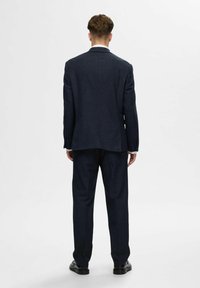 Marineblauer Anzug mit taillierter Passform, strukturiertem Stoff und einem Schlitz hinten. Kombiniert mit passender Hose und schwarzen Schuhen.