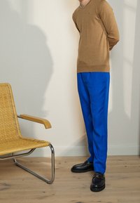 Pull en tricot marron, pantalon bleu vif, chaussures noires ; cadre comprenant une chaise en osier avec une structure chromée sur un sol en bois.