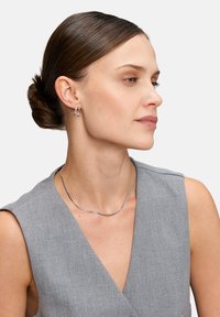 Eine Frau trägt silberne Creolen und eine glatte, gewölbte Silberkette zu einem grauen ärmellosen Oberteil mit V-Ausschnitt.