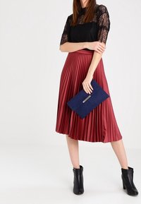 Černý krajkový top, plisovaná burgunská midi sukně, námořnická kabelka se zlatými detaily a černé kotníkové boty. Hladké textury a splývavý design.