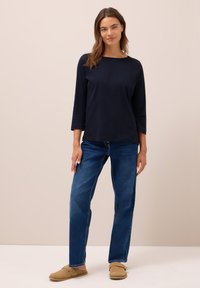 Femme se tenant contre un fond beige, portant un haut à manches longues bleu marine, un jean bleu et des chaussures de type slip-on beige.