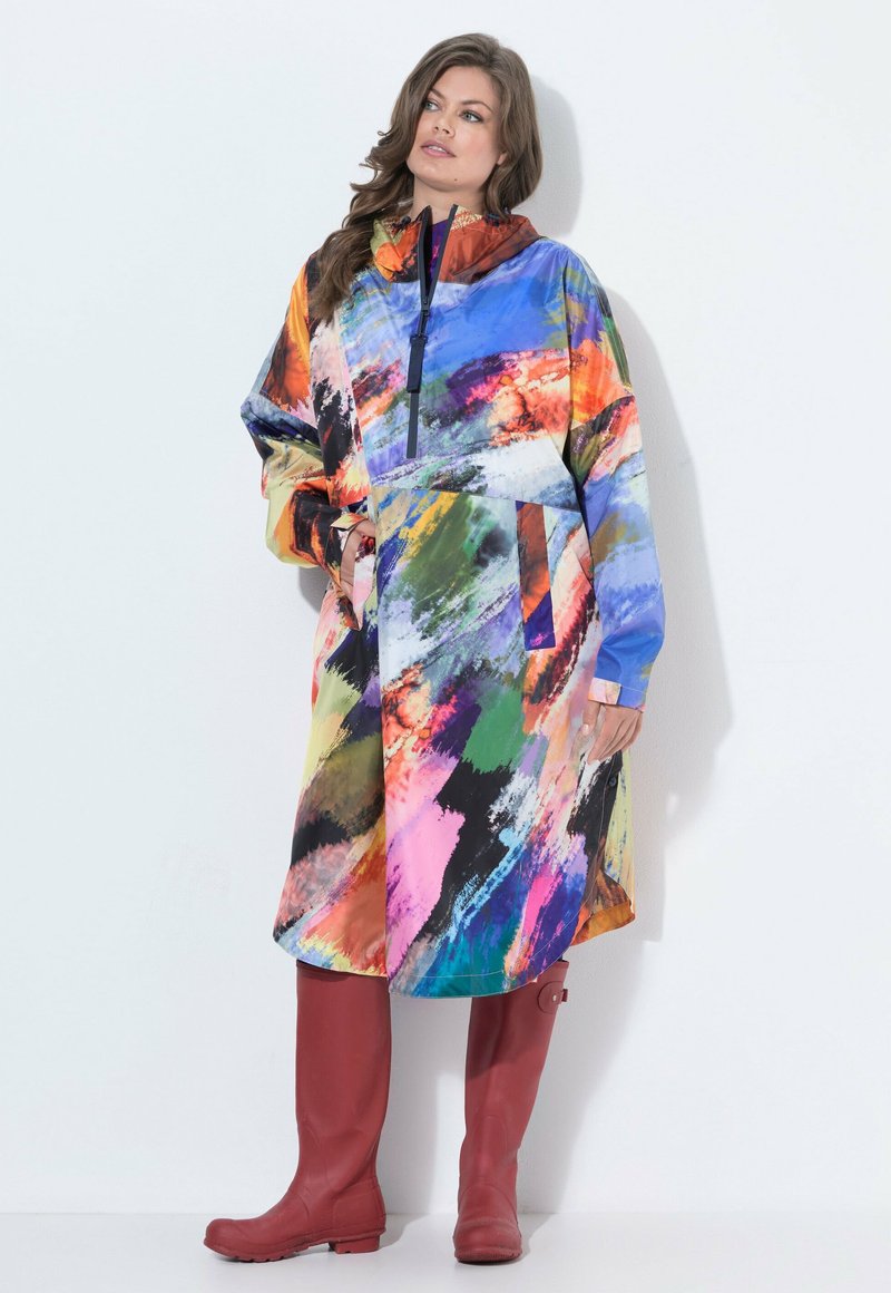 Woman wearing colorful abstract patterned raincoat with front zipper and red rubber boots standing against white background.