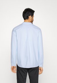 Camisa de manga larga azul claro con dobladillo curvado, puños con botones y un pliegue en la espalda que facilita el movimiento, fabricada con una tela suave.