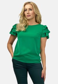 Zielona bluzka z krótkimi falbaniastymi rękawami, okrągłym dekoltem, gładką teksturą i luźnym krojem; zestawiona z ciemnozielonymi spodniami.