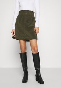 Vrouw in een olijfgroene corduroy rok met paperbag taille, witte langesleeve top en zwarte kniehoge haklaarzen op een effen achtergrond.