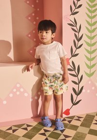 Kind draagt een lichtbeige T-shirt, geel met dieren bedrukt korte broek en blauwe rubberen sandalen, staand tegen een kleurrijke muur.