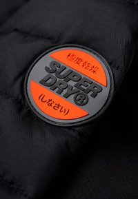 Schwarzer gesteppter Jackenstoff mit einem runden, schwarzen und orangefarbenen Gummilogo, auf dem "SUPER DRY®" steht, darunter japanischer Text.