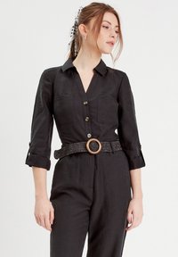 Camicia nera con colletto, due tasche sul petto, maniche arrotolate e una cintura intrecciata con fibbia circolare. Tessuto con una texture liscia.