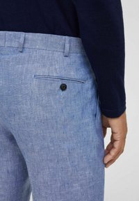 I pantaloni di lino azzurro chiaro presentano una tasca posteriore con un bottone nero e una texture sottile. Un maglione blu scuro è parzialmente visibile nella parte superiore.