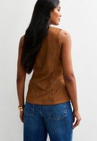 Top senza maniche in suede marrone con un design aderente, caratterizzato da una texture liscia e dettagli delle cuciture, abbinato a jeans blu a vita alta.