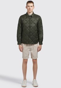 Donkergroene gewatteerde jas met een knoopsluiting en een puntige kraag, gecombineerd met beige shorts en witte sneakers. Geen zichtbare patronen of accenten.