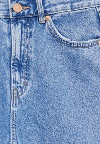 Gros plan d’un jean en denim bleu montrant la poche avant, la passants de ceinture, le bouton et les rivets métalliques avec des détails de couture visibles.