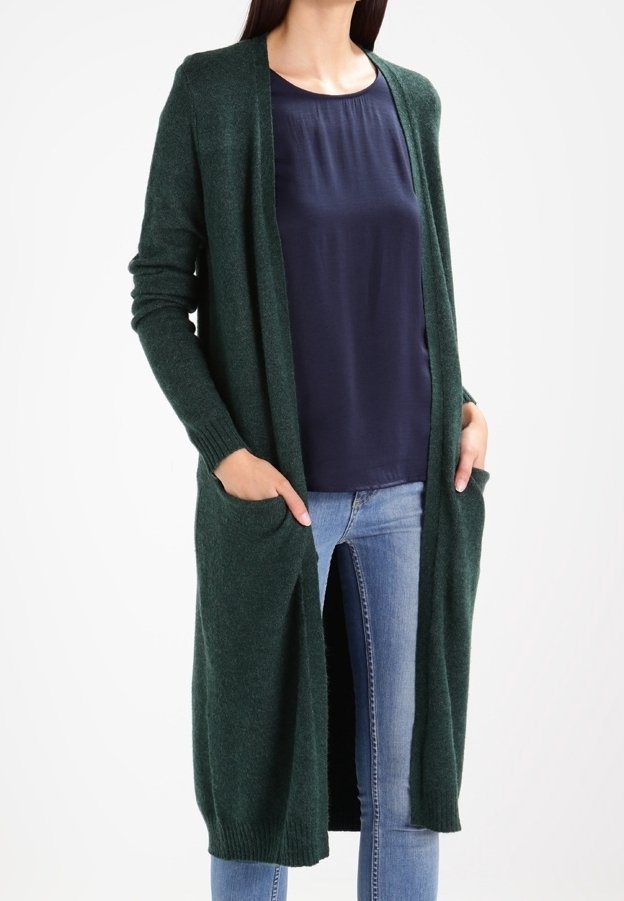 Femme portant un long cardigan ouvert vert foncé avec des poches, un haut bleu marine et un jean bleu clair, debout avec les mains dans les poches.