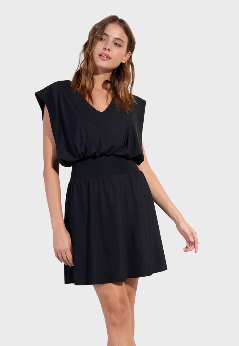 Femme aux cheveux bruns mi-longs portant une robe noire sans manches avec un col en V et une taille cintrée, debout devant un fond uni.