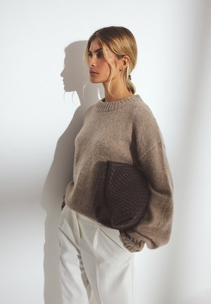 Beige strikket sweater med ribbet halsudskæring, parret med hvide bukser. En mørk vævet clutch holdes under hendes arm mod en lys baggrund.