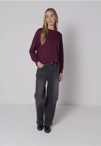 Camicia bordeaux a maniche lunghe con scollo rotondo, abbinata a jeans neri larghi e stivali neri. Tessuto morbido, vestibilità comoda, look casual.