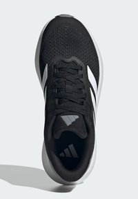 Scarpa da ginnastica atletica bianca e nera con tomaia in mesh, lacci e logo Adidas sulla linguetta e sulla soletta, vista dall'alto su uno sfondo chiaro.