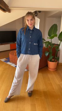 Džinsinė marškinė su ilgomis rankovėmis, sutrumpintu dizainu ir baltu logotipu. Pora su laisvomis, šviesiomis lino kelnėmis ir pilkais batais.