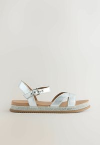 Sudraba krāsas plakanas sandales ar potītes sprādzes siksnu un rhinestone rotātu zoli uz vienkārša gaiša fona.