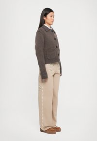 Cardigan marrone lavorato a maglia con grandi bottoni e colletto, abbinato a pantaloni beige ricamati. Presenta tasche laterali e dettagli testurizzati.
