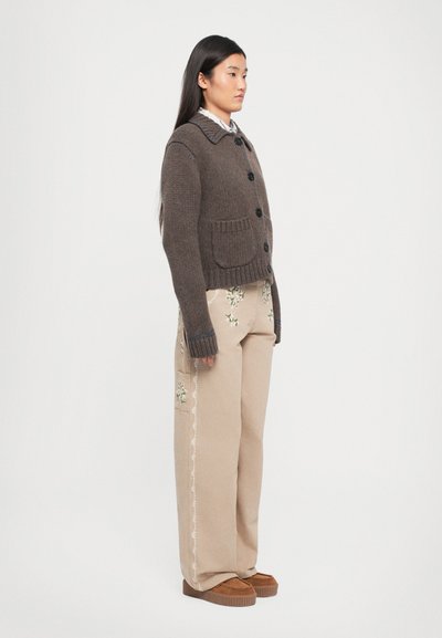 Braune Strickjacke mit großen Knöpfen und einem Kragen, kombiniert mit beigen bestickten Hosen. Verfügt über seitliche Taschen und strukturierte Akzente.