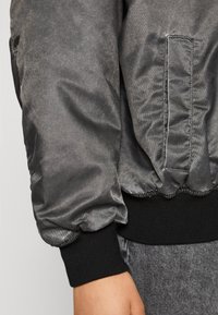 Giacca bomber grigia in satin con polsini elasticizzati, chiusura con zip e tasca laterale, caratterizzata da una texture liscia e un finish leggermente lucido.