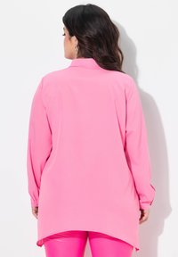 Langärmlige Bluse aus leuchtend pinkem Stoff, mit Kragen und geschwungenem Saum, kombiniert mit glänzenden pinken Leggings. Glatte Textur.