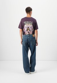 La camiseta púrpura presenta un estampado gráfico colorido en la parte posterior. El modelo lleva jeans rectos de color azul claro y zapatillas blancas.
