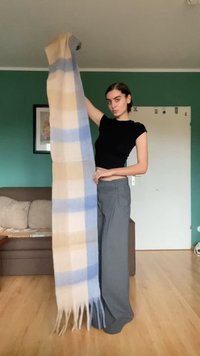 Langer Schal in hellbeige mit blauen Streifen, aus weichem Stoff gefertigt, mit fransigen Enden. Model hält ihn vertikal.