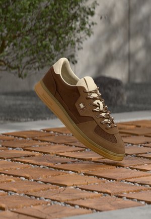 Scarpa da ginnastica marrone e beige con suola testurizzata e lacci, sospesa sopra un pavimento esterno in legno con motivi e ciottoli, vicino a piante e a un muro.