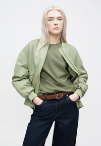 Ελιά πράσινη bomber jacket με ριχτούς μανίκια και λαιμόκοψη, συνδυασμένη με ένα ταιριαστό t-shirt και σκούρα τζην παντελόνια, συγκρατημένα με καφέ ζώνη.