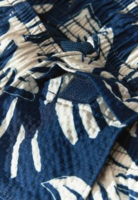 A sötétkék textil felszíne texturált mintázattal rendelkezik, fehér levélmintákkal, kontrasztos hálós kiemelésekkel és összehúzott varrásokkal, hogy még több dimenziót adjon.