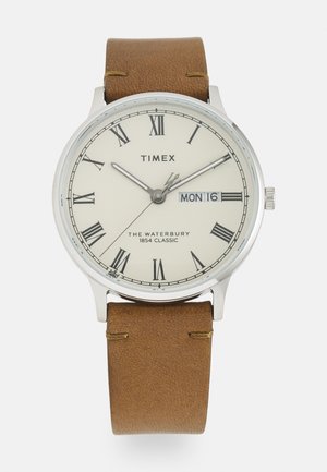 Montre ronde en argent avec un cadran blanc et des chiffres romains, bracelet en cuir marron et affichage de la date pour le lundi 16.