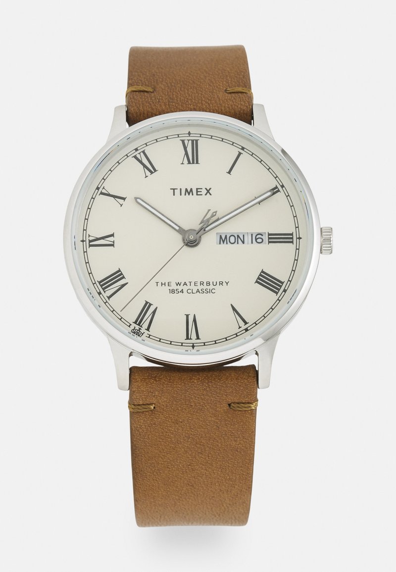 Reloj de plata redondo con un dial blanco con marcadores en números romanos, correa de cuero marrón y una pantalla de fecha para el lunes 16.