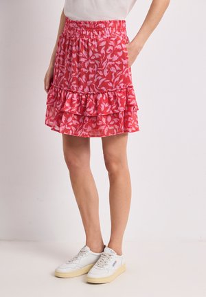 Femme portant une mini-jupe à volants à motif floral rouge associée à des baskets blanches, debout avec une main dans la poche.