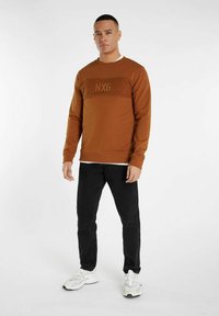 Bruine sweatshirt met textuurvolle NXG-logo op de borst, gecombineerd met zwarte jeans en witte sneakers. Glad stof met een ontspannen pasvorm.