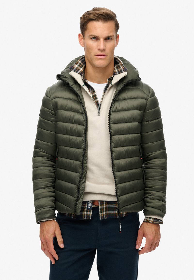 Polstret mørkegrøn jakke med lynlås forsejling og hætte, der har et ternet mønster på kraven, båret over en lysefarvet sweater.
