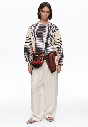 Młoda kobieta nosząca pasiastą bluzkę z długim rękawem, szerokie białe spodnie, klapki, z czerwonym swetrem związanym w talii oraz plecioną torbą na ramię.