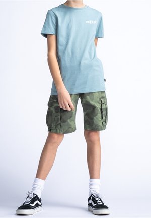 Camiseta de algodón azul claro con estampado de "PETRI", combinada con pantalones cortos cargo de camuflaje verde con bolsillos laterales. Zapatillas negras con detalles blancos.
