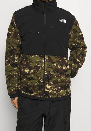 Mężczyzna ubrany w czarną i zieloną kamizelkę polarową w kamuflażu z logo The North Face na piersi, czarne spodnie i z jedną ręką w kieszeni.