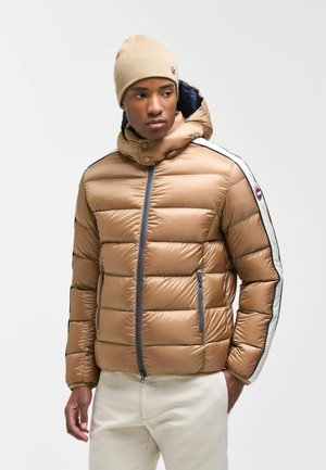 Giovane uomo che indossa una giacca imbottita beige con strisce bianche e nere sulle maniche e un berretto beige abbinato, in piedi davanti a uno sfondo semplice.