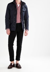 Veste bleu marine, imperméable, avec plusieurs poches, portée sur une chemise rayée rouge. Un pantalon noir ajusté et des mocassins marron foncé complètent le look.