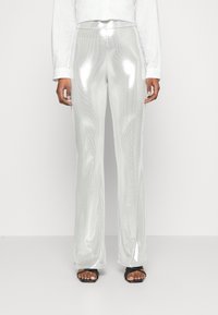 Person wearing shiny silver pants, white long-sleeve shirt, and black open-toe sandals, standing against a plain white background.