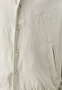 Veste gris clair en tissu doux, avec une fermeture à boutons et des poches latérales. Texte brodé "New York City 1990" visible.