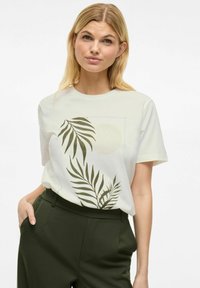 Valkoinen lyhythihainen t-paita, jossa on vihreä palmunlehden kuvio ja hillitty pyöreä muotoilu. Yhdistetty tummanvihreisiin korkeavyötäröisiin housuihin.