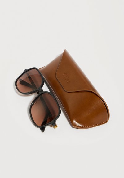 Καφέ φιμέ γυαλιά ηλίου με μαύρο σκελετό δίπλα σε καφέ δερμάτινη θήκη με κλείσιμο με καπάκι, σε λευκό φόντο.