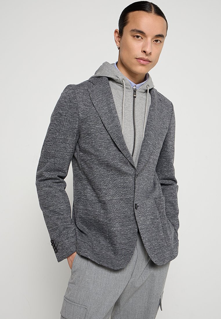 Szary, teksturowany żakiet nałożony na jasnoszary, bluzę z kapturem, z pojedynczym zapięciem na guzik i dwoma przednimi kieszeniami.